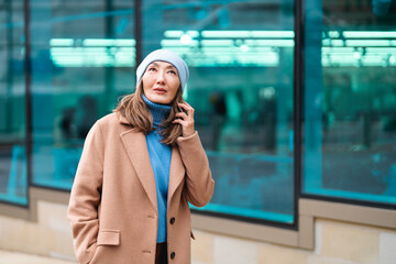 Fototapeta premium Woman in a beige coat and blue beanie looks up thoughtfully while talking on her phone in an urban setting