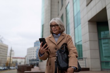 Senior woman using smartphone while standing outside a modern building on a cloudy day in the city