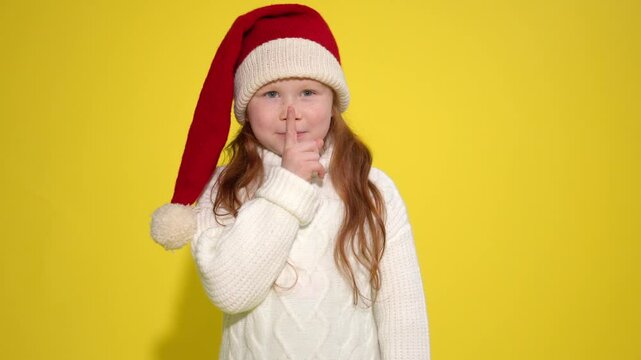 Cute child wearing festive hat holding finger to lips in shush gesture in bright yellow background during holiday season