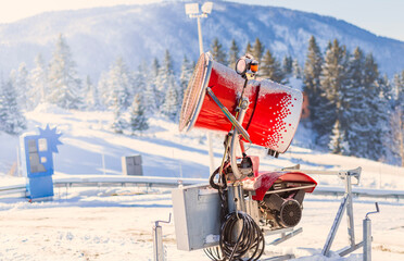 "Snow cannon machine producing artificial snow at a ski resort on a sunny winter day. Snow-covered mountains and evergreen trees in the background create a picturesque alpine scene."