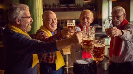 Elderly people, men in colorful red and yellow scarves meeting at bar, pub, clinking beer mugs and celebrating victory of favorite soccer team. Sport fans, elderly lifestyle, leisure activity concept