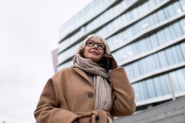 Elderly woman smiling while talking on the phone outdoors near modern building on a cloudy day