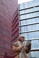 A woman uses a tablet while standing in front of a modern glass building with red accents during a...