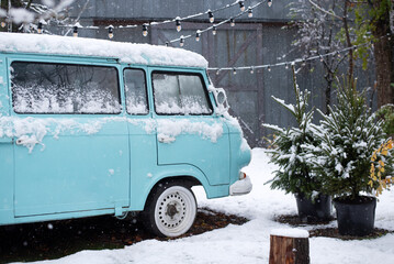 Retro blue van covered in snow, parked near pine trees and string lights in a snowy winter setting