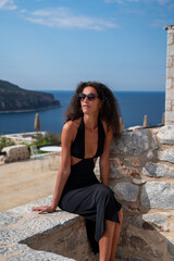 Fototapeta premium Woman model with curly hair in a black dress enjoying and black sunglasses the seaside view in Greece