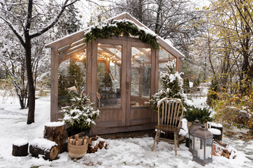 Cozy wooden greenhouse decorated with Christmas lights, trees, and snow in a winter garden setting