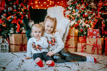 Two young girls in matching festive outfits sitting by a Christmas tree surrounded by presents,...