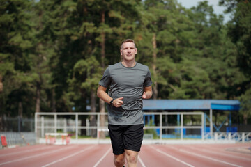 Man jogging on a track surrounded by trees, wearing grey shirt and black shorts. Background includes outdoor sports facility and lush greenery