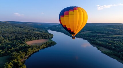 Obraz premium A majestic yellow hot air balloon peacefully glides above a lush, river valley, surrounded by expansive forested hills under a clear blue sky, inspiring awe.
