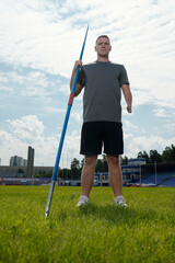 Athlete holding javelin on sports field with stadium and sky in background, demonstrating...