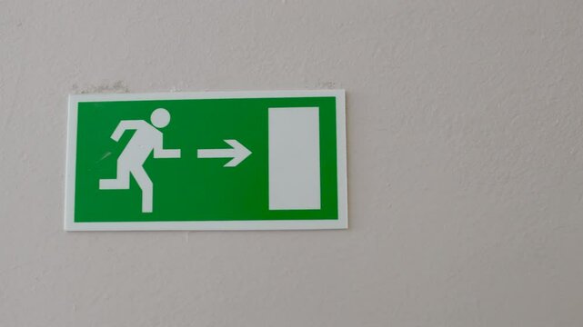 Green emergency exit sign with white running figure and arrow pointing towards door. Indicating direction for quick and safe evacuation route in case of emergency.