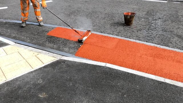 Road worker applies bright redb coating to pavement in urban area during daytime