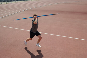 Athlete running on red track field holding javelin above shoulder and preparing release. Background includes part of stadium and fencing surrounding track field