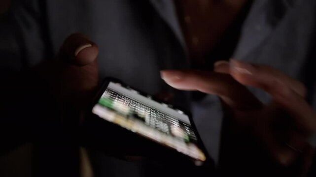 Close-up of hands scrolling through a newsfeed on a smartphone in a dimly lit setting. The light from the screen illuminates the fingers as they navigate the content. Perfect for projects related to