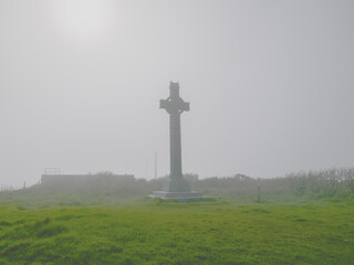 A tall Celtic cross is prominently displayed in a lush green field, surrounded by thick fog in the early morning. The atmosphere is tranquil and mystical, creating an ethereal setting.