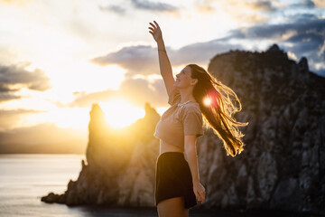 A young woman dancing at sunset among the sea and mountains background. Golden sunbeams shine on a beautiful girl twirling. Feeling of freedom at sunset. Travel. Adventure. Vacation. Summer holidays.