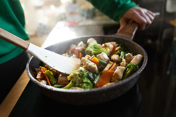 A woman frying fresh healthy vegetables and chicken in a pan on an induction stove in a home...