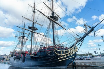 Historic sailing ship docked at a harbor under a bright blue sky with fluffy clouds