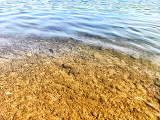 Sandy bottom is visible through clear water near the shore of a tranquil lake on a sunny afternoon