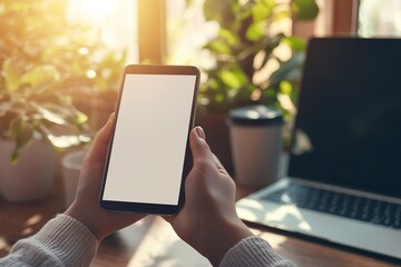 Hands holding a smartphone with a blank screen in a sunlit home workspace