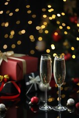 Delicious champagne in glasses and Christmas decor on black mirror surface against blurred lights. Space for text