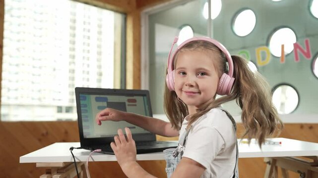 Smiling girl using laptop and turn around to show thumb up to camera. Cute child wearing headphone while working by using laptop writing code in STEM technology education. Online learning. Erudition.
