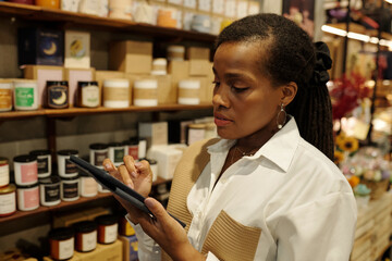 African American woman holding tablet while browsing through items displayed on shelves in store. Paying close attention to selecting products, creating a focused atmosphere © DragonImages