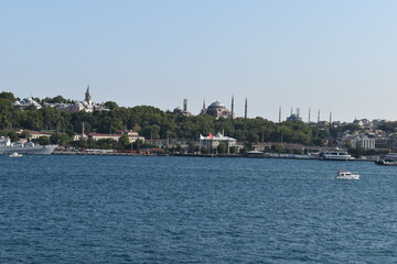 Fototapeta premium A panoramic view of Istanbul's historical peninsula from the Bosphorus Strait, showcasing the Topkapi Palace and Hagia Sophia, surrounded by lush greenery. A small boat navigates the water in the fore
