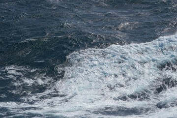 Waves crashing on rocky shore, creating foam and ripples in ocean. deep blue water contrasts with white froth, evoking sense of tranquility and power
