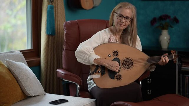 Mature woman sits in a leather chair, holding a traditional oud guitar and looking at her smartphone. She is wearing a white blouse and black pants, with long blonde hair. The room is decorated