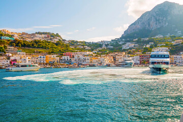 Fototapeta premium The photo captures a view of Capri from the water, showcasing the island's iconic cliffs, colorful buildings, and lush greenery set against the clear blue sea and sky.