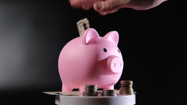 Male hand inserts a banknote into a pink piggy bank surrounded by scattered coins and additional banknotes, creating a vivid contrast against the dark background