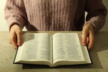Woman reading Holy Bible in English language at beige table, closeup
