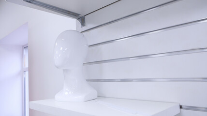 White acrylic mannequin head mounted on a white shelf. The mannequin is placed against the background of a panel with metal guides intended for installing commercial equipment.