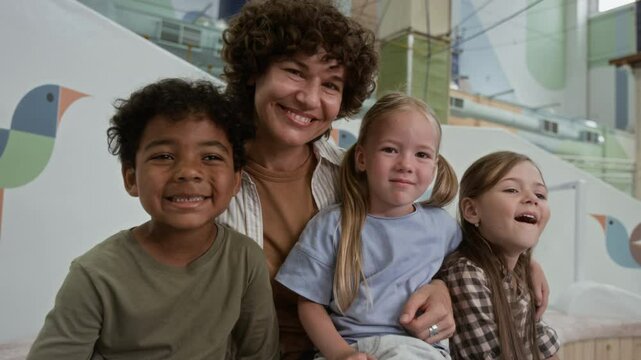 Low angle portrait of cheerful Caucasian female animator with curly hair cuddling diverse gen Alpha kids and smiling at camera in childrens room