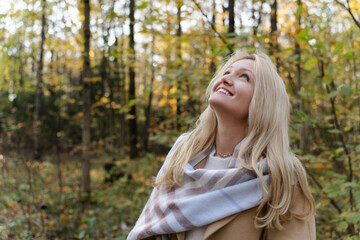 young dreamy woman with blonde hair smiles brightly while walking in a peaceful autumn park. The warm sunlight and golden leaves create a cozy, serene atmosphere