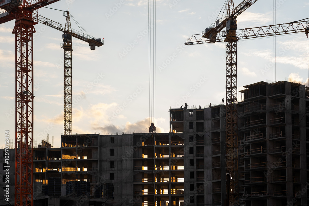 Poster cranes lift materials as workers construct a large building at a construction site illuminated by a 