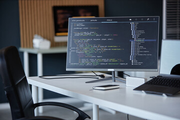 Image of a modern office workspace featuring a computer monitor displaying software code and various desk items. Background includes blurred office setup with chairs and shelves seen