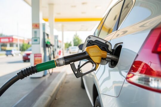 A silver car is being refueled at a gas station during the daytime. The fuel nozzle is inserted into the vehicle's tank as the driver prepares to fill up the gas tank. Nearby cars are parked, adding t