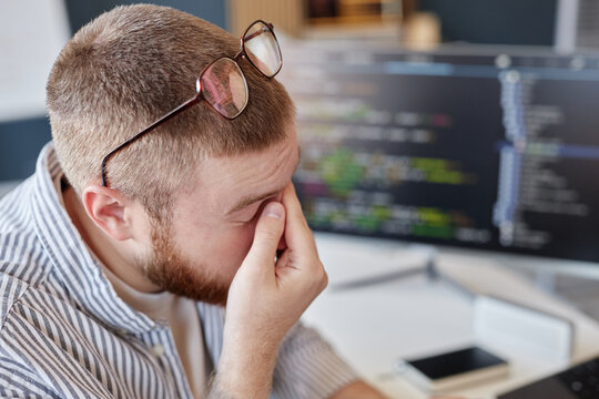 Male software developer rubbing eyes while sitting at desk in front of computer screen with code. Glasses resting on top of head, showing signs of discomfort and fatigue