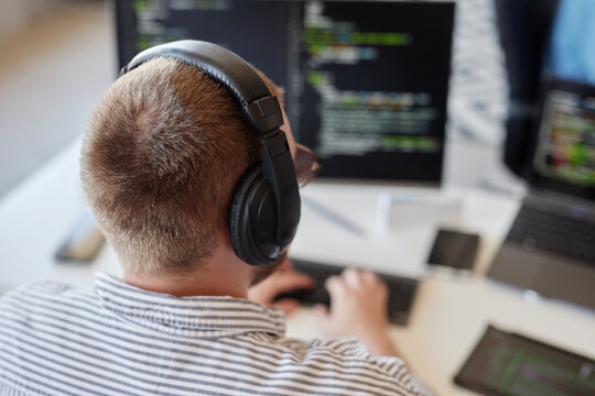 Person sitting at desk and wearing black over-ear headphones while focusing on coding on computer screen with code in background