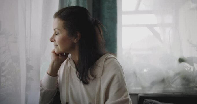 Woman gazing thoughtfully out of window, resting chin on hand, sitting in cozy home setting with natural light, reflecting in serene, calm environment