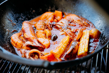 Seafood boil in a wok with shrimp, squid, corn, and rich sauce, cooking over a grill