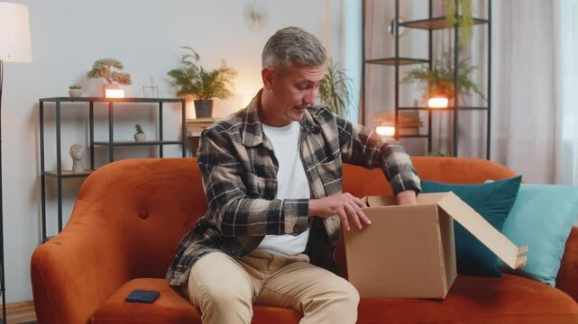 Unhappy young Caucasian man guy unpacking carton box, feeling dissatisfied with getting damaged wireless headphones order, writing negative feedback in mobile application, internet shopping concept.