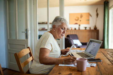 Concerned woman reading bills on kitchen table with laptop