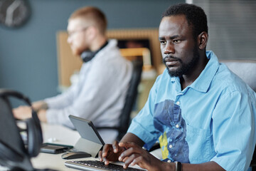 African American man working on laptop in modern office setting filled with light emitting a calm focus, while a colleague is seen in background engaged with his tasks