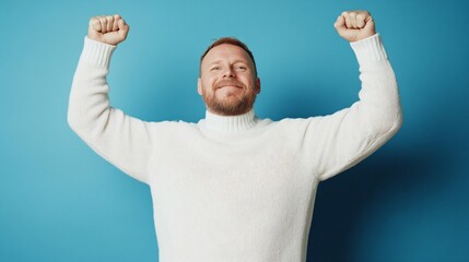 Fototapeta premium Happy person in white sweater celebrating success with raised arms against blue background. Joyful victory pose. Winning gesture. Positive emotion concept for motivation and achievement