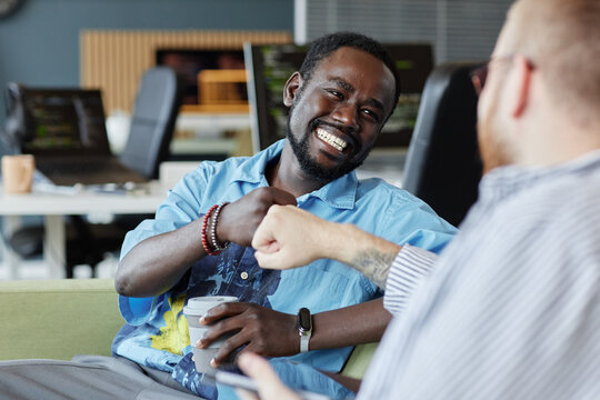 Smiling African American man fist-bumping with colleague while holding coffee cup in modern office setting, displaying camaraderie and friendly teamwork