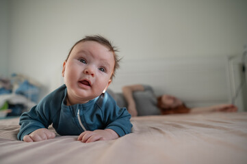 A three-month-old boy lies on his stomach on the bed and his mother sleeps behind him. Postpartum...