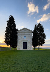 A stone chapel stands on a grassy hill at sunset, surrounded by tall trees